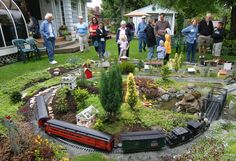 a group of people standing around a model train set in a yard with trees and bushes