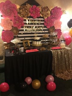 a table topped with balloons and cake next to a wall covered in paper flower decorations