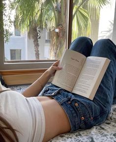 a woman laying on her stomach reading a book in front of a window with palm trees