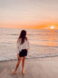 a woman standing on the beach at sunset with her back to the camera, facing the water