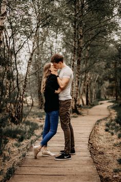 a man and woman standing on a wooden walkway in the woods hugging each other with trees behind them