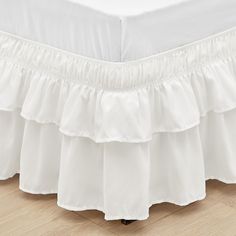 a white bed skirt with ruffles on the bottom and bottom layer, sitting on top of a wooden floor