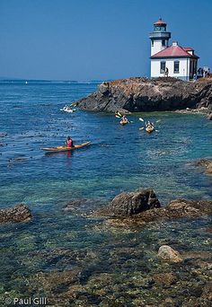 people are paddling their canoes in the clear blue water near a light house