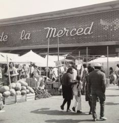 black and white photograph of people walking in front of a market with produce on display