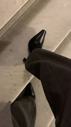 a person wearing black high heel shoes standing next to a white wall and stairs with their feet on the ground