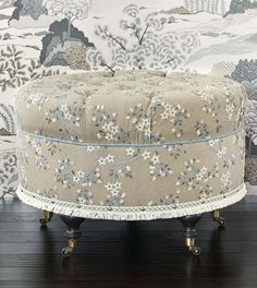 an upholstered ottoman sits in front of a wallpapered background with flowers