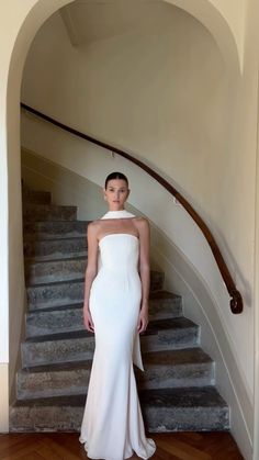 a woman in a white dress standing at the bottom of stairs with her hands on her hips