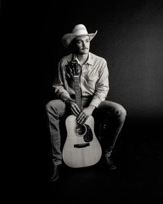 a man with a cowboy hat sitting on a chair holding an acoustic guitar in his hands