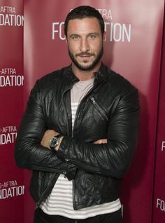 a man with his arms crossed standing in front of a red wall wearing a black leather jacket