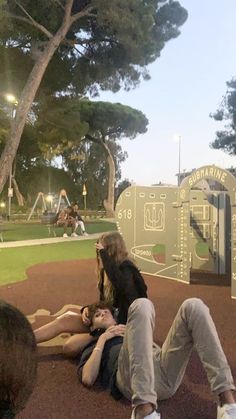 three people sitting on the ground in front of a playground with trees and lights behind them