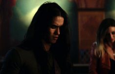 a man standing next to a woman with long hair and wearing a black jacket in a dark room