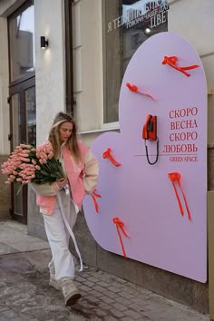 a woman is walking down the street with flowers in her hand and a pink heart shaped sign behind her