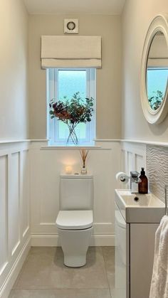 a white toilet sitting next to a sink in a bathroom under a window with a flower vase on it