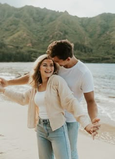 a man and woman standing on the beach holding hands, smiling at each other with mountains in the background