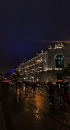 people walking down the street at night in front of a large building with lights on it