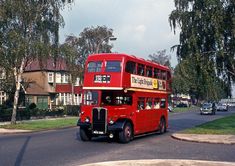 a red double decker bus driving down a street next to tall buildings and trees in the background