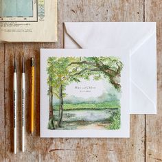 a card with watercolors and pencils next to it on a wooden table