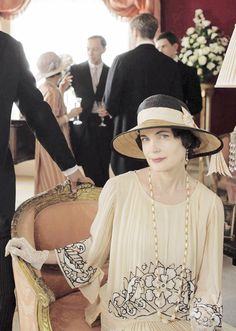 a woman in a dress and hat sitting on a chair next to a man in a suit