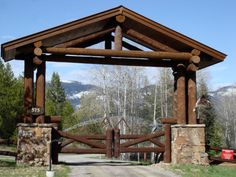 a wooden gate with stone pillars in the middle of a dirt road surrounded by trees and mountains