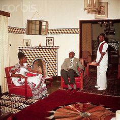 two men and a woman sitting in chairs in a living room next to a fire place