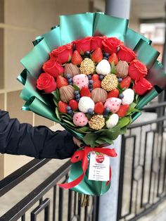 a person holding a bouquet of flowers with chocolates and strawberries in it on a railing