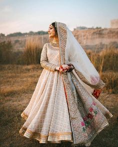 a woman in a white and gold lehenga standing in the grass with her hands on her hips