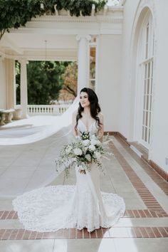 a woman standing in front of a white building holding a bouquet and wearing a wedding dress