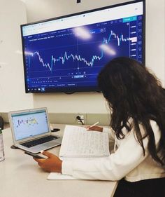 a woman sitting at a desk in front of a laptop computer with a monitor on the wall behind her