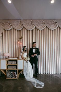 a bride and groom standing next to each other in front of a wall with curtains