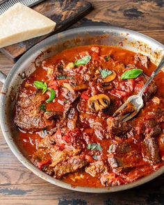 a skillet filled with meat and tomato sauce on top of a wooden table next to bread