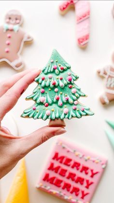 a hand holding a decorated christmas tree cookie next to cookies and other holiday decorations on a table