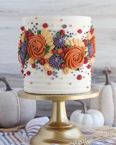 a white cake with orange and purple flowers sitting on top of a gold stand next to pumpkins