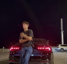 a man leaning on the hood of a black car at night with his arms crossed