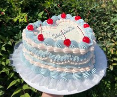 a blue and white cake with cherries on top sitting in front of some bushes