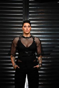 a woman standing in front of a metal wall wearing black pants and a sheer top