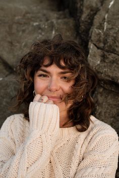 a woman with curly hair wearing a white sweater posing for a photo in front of some rocks