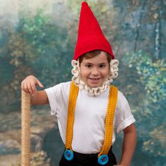 a young boy wearing a red gnome hat and suspenders holding a stick in front of a painting