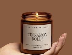 a hand holding a candle that says cinnamon rolls in front of a dark brown background