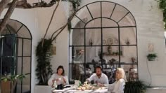 three people sitting at a table outside in front of a building with large arched windows