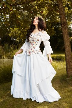 a woman wearing a white dress standing in front of a tree with her hands on her hips