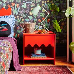 a bedroom with colorful wallpaper and a red nightstand next to a green plant in the corner