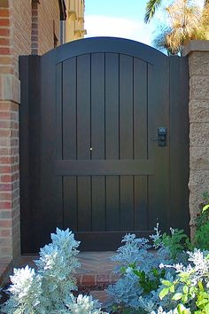 a large wooden gate in front of a brick building with blue flowers and greenery