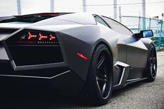 the rear end of a black sports car parked in front of a fenced area