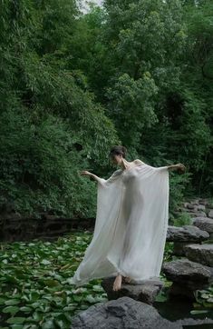 a woman is standing on rocks in the water with her arms spread out and she is wearing a flowing white dress