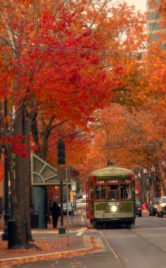 a green bus driving down a street next to trees with red and orange leaves on it