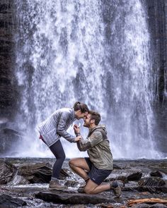 a man kneeling down next to a woman in front of a waterfall