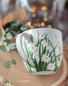 a coffee cup with daisies painted on the side and green leaves in the background