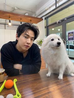 a man sitting at a table next to a white dog on top of a wooden floor