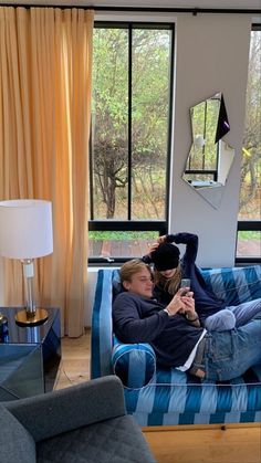 two people sitting on a blue couch in front of a window looking at their cell phones