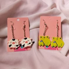 two pairs of earrings with cows on them sitting on a pink sheet covered bed next to each other